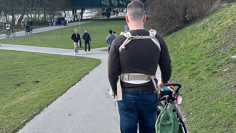 Simon Picht schiebt den Rollstuhl seines Sohnes und trägt sein Baby vor dem Bauch.