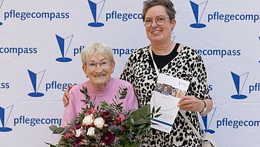 Zwei Frauen stehen vor einer „pflegecompass-Fotowand“, die rechte Person hat eine Urkunde in der Hand und legt den Arm um eine ältere Frau, die einen Blumenstrauß in der Hand hat. Beide lächeln in die Kamera. 