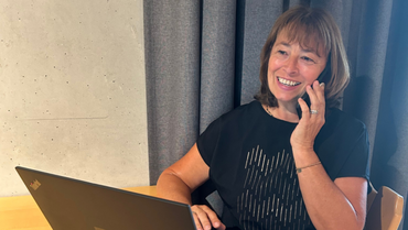 Eine Frau sitzt an einem Tisch. Der Laptop steht aufgeklappt vor ihr, sie telefoniert und lächelt.