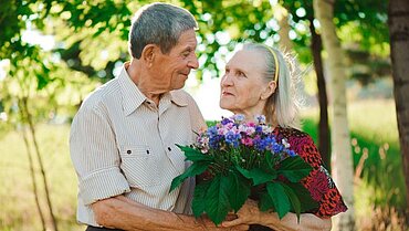 Das Bild zeigt ein älteres Paar im Grünen, das sich zugewandt ist. Die Frau hält einen Blumenstrauß aus Wildblumen.