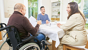 Ein Pflegeberater spricht an einem Tisch sitzend mit einem pflegebedürftigen Mann im Rollstuhl und dessen Ehefrau.