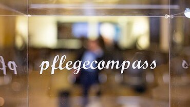Die transparente Losbox des pflegecompass, im Hintergrund Menschen.