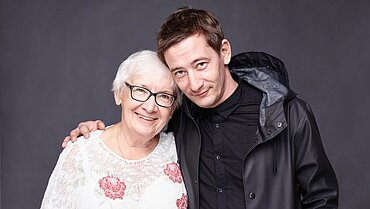 Eine ältere Frau und ein Mann mittleren Alters stehen lächelnd nebeneinander und haben jeweils einen Arm um die andere Person gelegt.