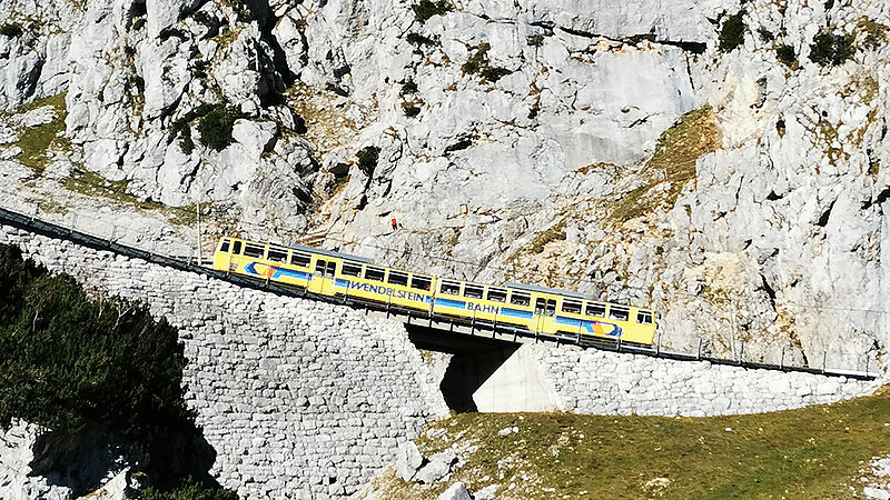 Eine mit Pflanzen spärlich bewachsene Gebirgswand, vor der eine gelbe Bahn entlangfährt.