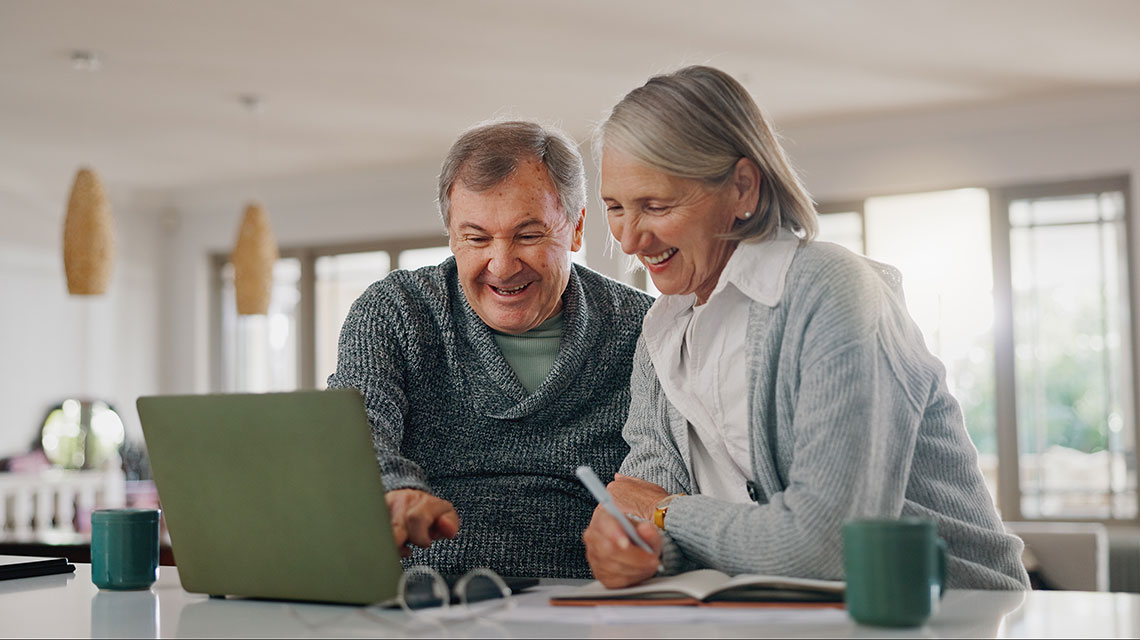 Ein Mann und eine Frau, die beide älter sind, schauen gemeinsam auf einen Computer. Sie lächeln und machen sich Notizen.