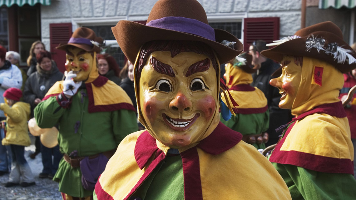 Mehrere Personen, welche traditionelle Gewänder und handgefertigte Masken tragen, ziehen durch die Straße. 