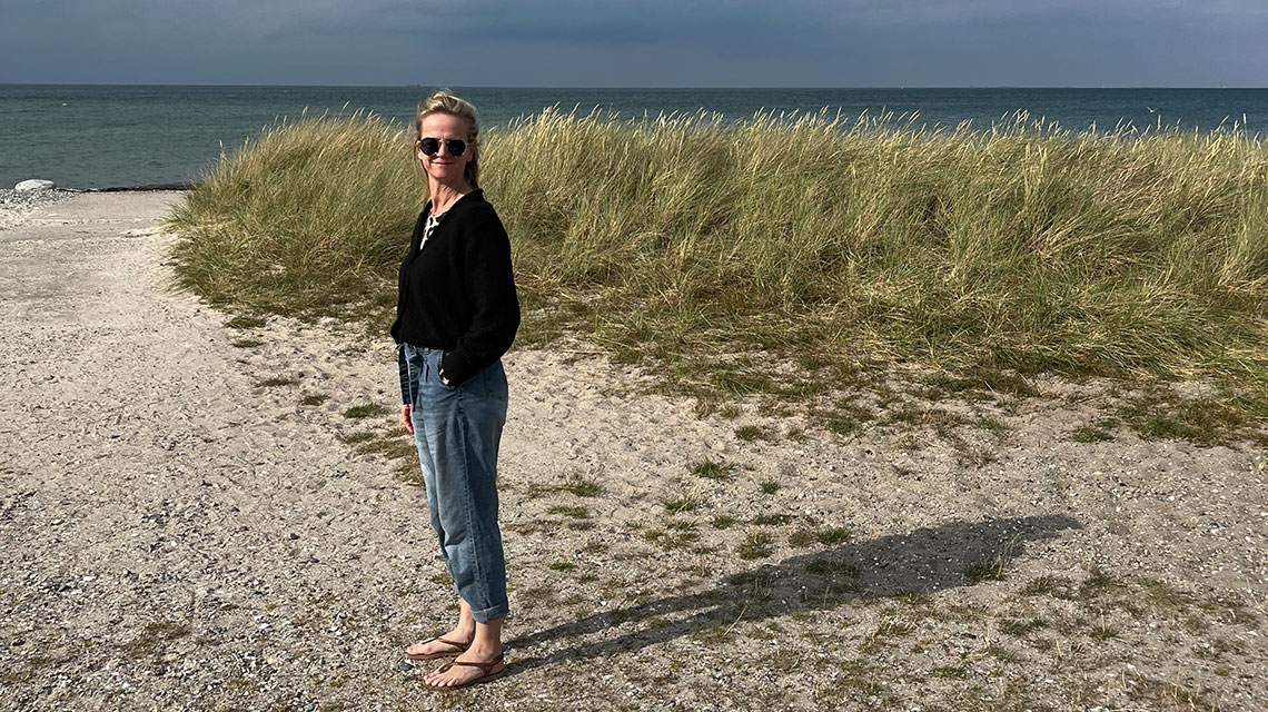 Nicole Zahn steht mit den Händen in der Hosentasche auf einem Sandabschnitt und schaut in die Kamera, im Hintergrund ist das Meer und ein teils bewölkter Himmel. 