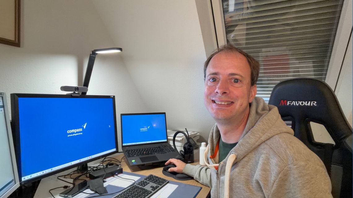 Ein Mann sitzt an einem Schreibtisch vor einem Laptop und einem Bildschirm und lächelt in die Kamera.