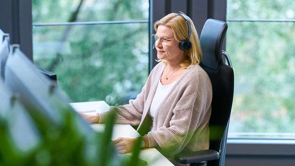 Pflegeberaterin sitzt mit Headset auf einem Bürostuhl vor dem Computer.