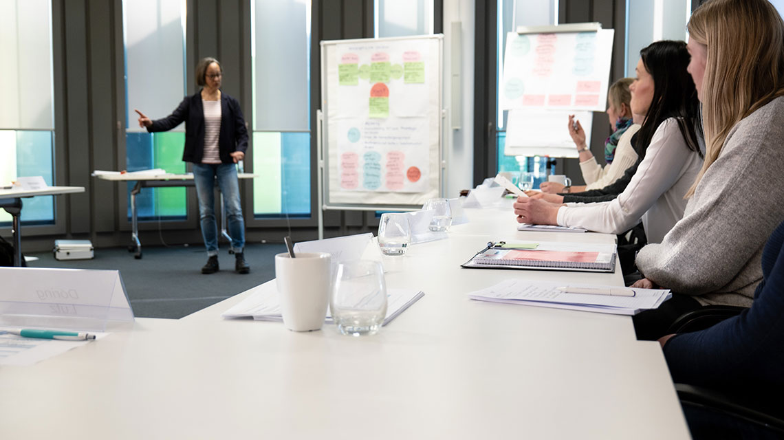 Eine Gruppe neuer Kolleg*innen nimmt am Unterricht in einem Konferenzraum teil. Eine Dozentin steht vorne.