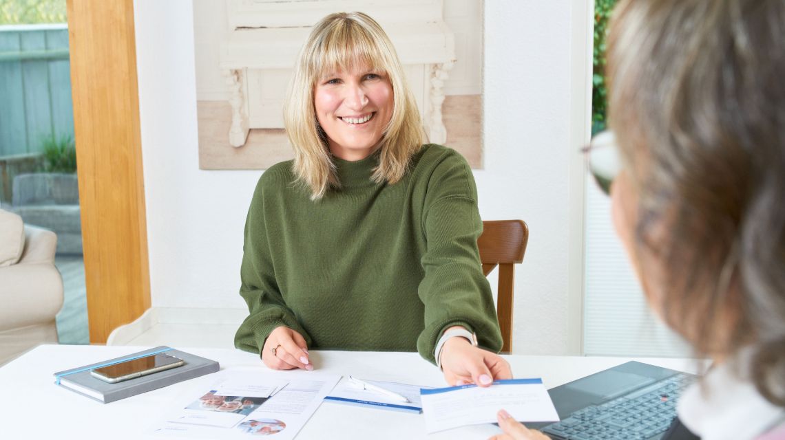 Eine Pflegeberaterin sitzt mit einer Frau am Tisch und gibt ihr eine Unterlage. Die Pflegeberaterin lächelt.