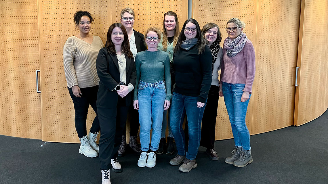 Acht Frauen in legerer Kleidung stehen nebeneinander und lächeln in die Kamera. Sie befinden sich in einem Raum, der Hintergrund ist eine Schrankwand.