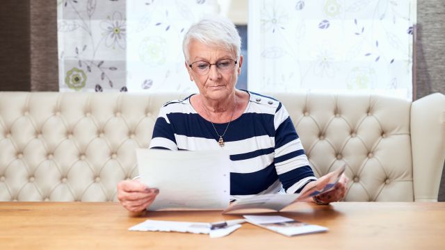 Seniorin mit kurzen Haaren sitzt an Tisch vor einem Fenster und sieht Dokumente durch.