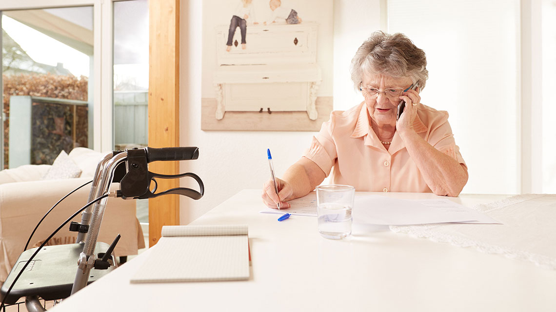Eine ältere Dame sitzt telefonierend an einem Tisch und macht sich Notizen auf einen Block. Nebenan steht ein Rollator.
