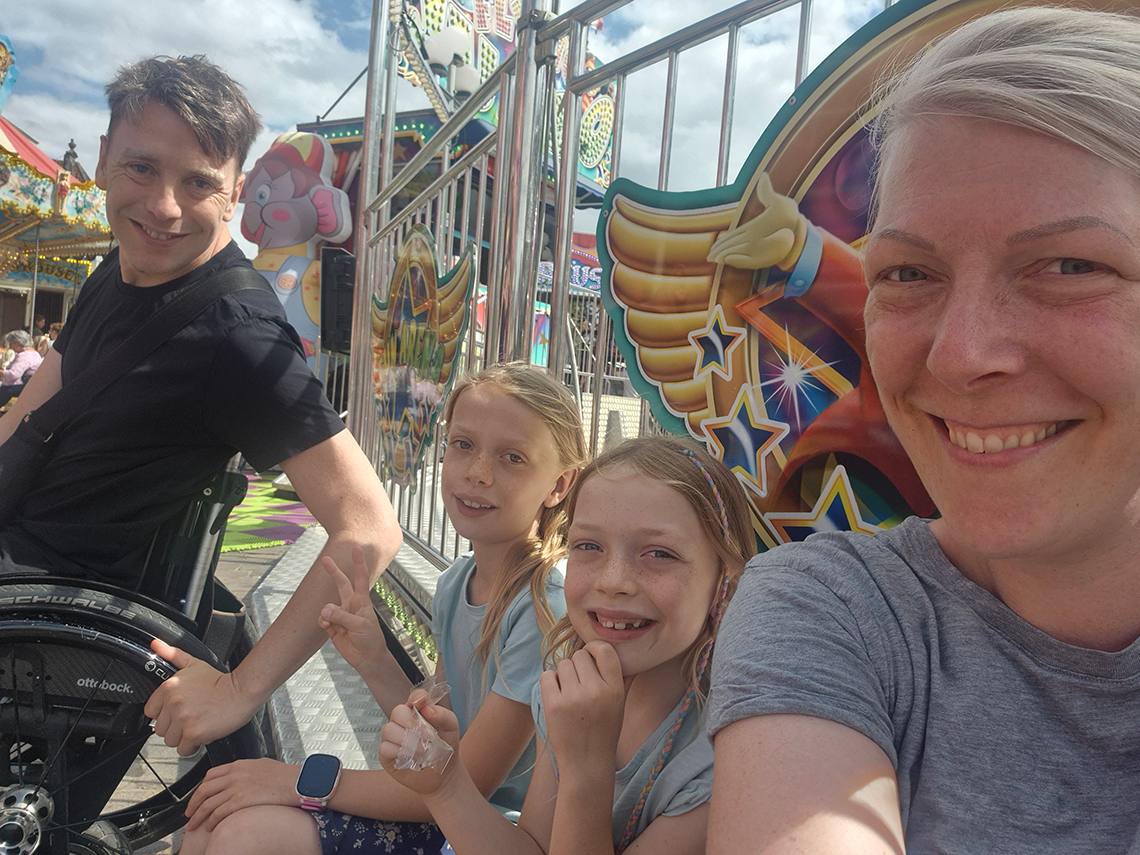 Ein erwachsener Mann im Rollstuhl, zwei Kinder und eine erwachsene Frau lachen in die Kamera. Im Hintergrund sieht man eine Kirmes.
