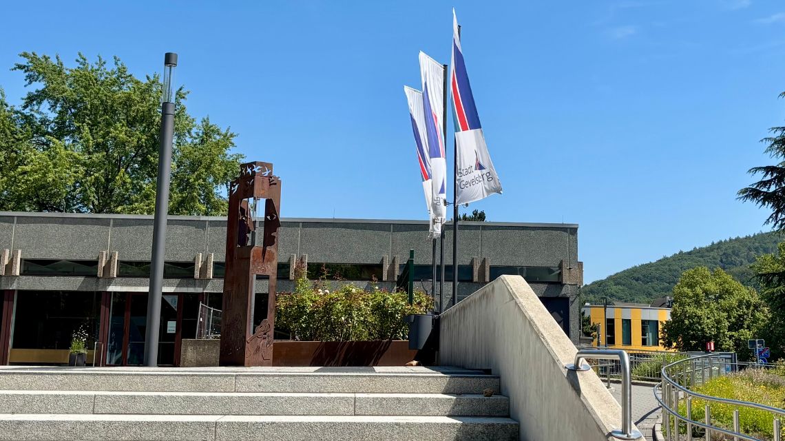 Ein graues Gebäude mit Treppenaufgang und drei Fahnen mit der Aufschrift „Stadt Gevelsberg“ bei blauem Himmel. 	