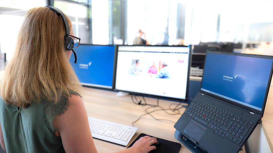 compass-Beraterin sitzt mit Headset an ihrem Schreibtisch und blickt freundlich in die Kamera.