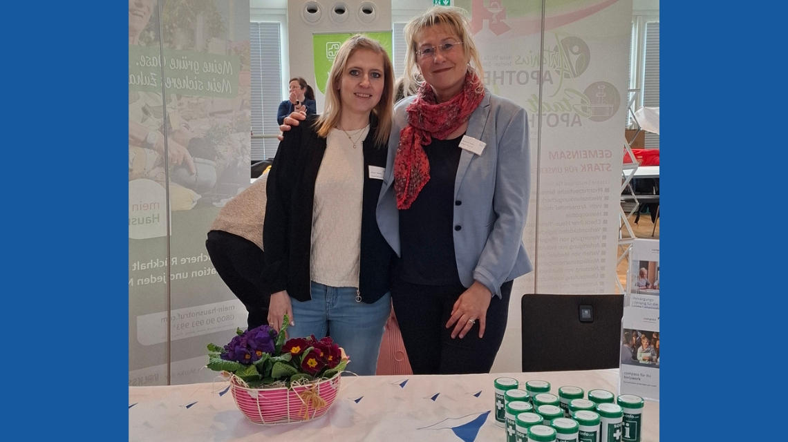 Mandy Böttger und Simone Schumacher stehen an einem Messestand Arm in Arm und lächeln in die Kamera.  