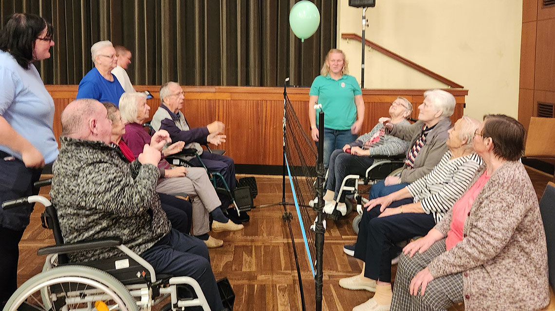 Mehrere ältere Menschen sitzen in einer Sporthalle auf zwei Seiten, getrennt von einem Netz und halten einen Luftballon in der Höhe.  