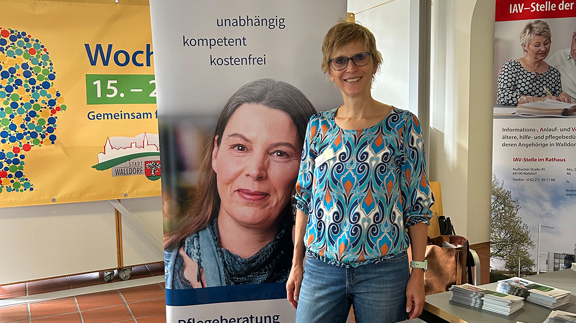 Frau steht vor einem Banner und lächelt in die Kamera. Vor ihr liegen Prospekte auf einem Tisch. 