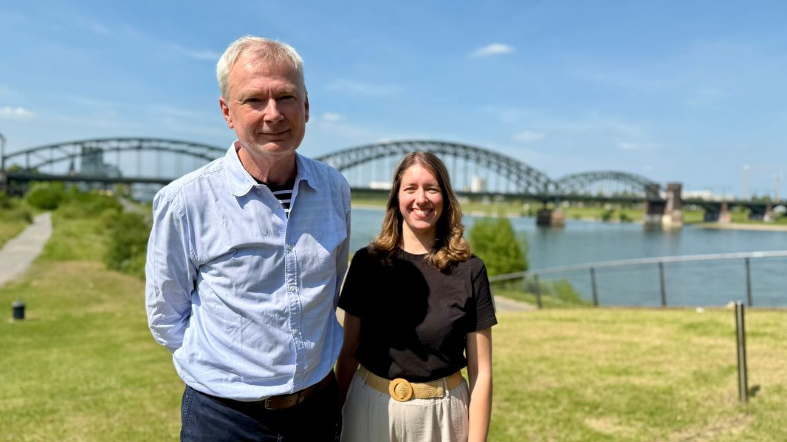 Olaf Arps (links) und Leonie Hausmann (rechts) nebeneinander am Rhein in Köln. Es ist ein sonniger Tag. Im Hintergrund ist die Südbrücke zu sehen.